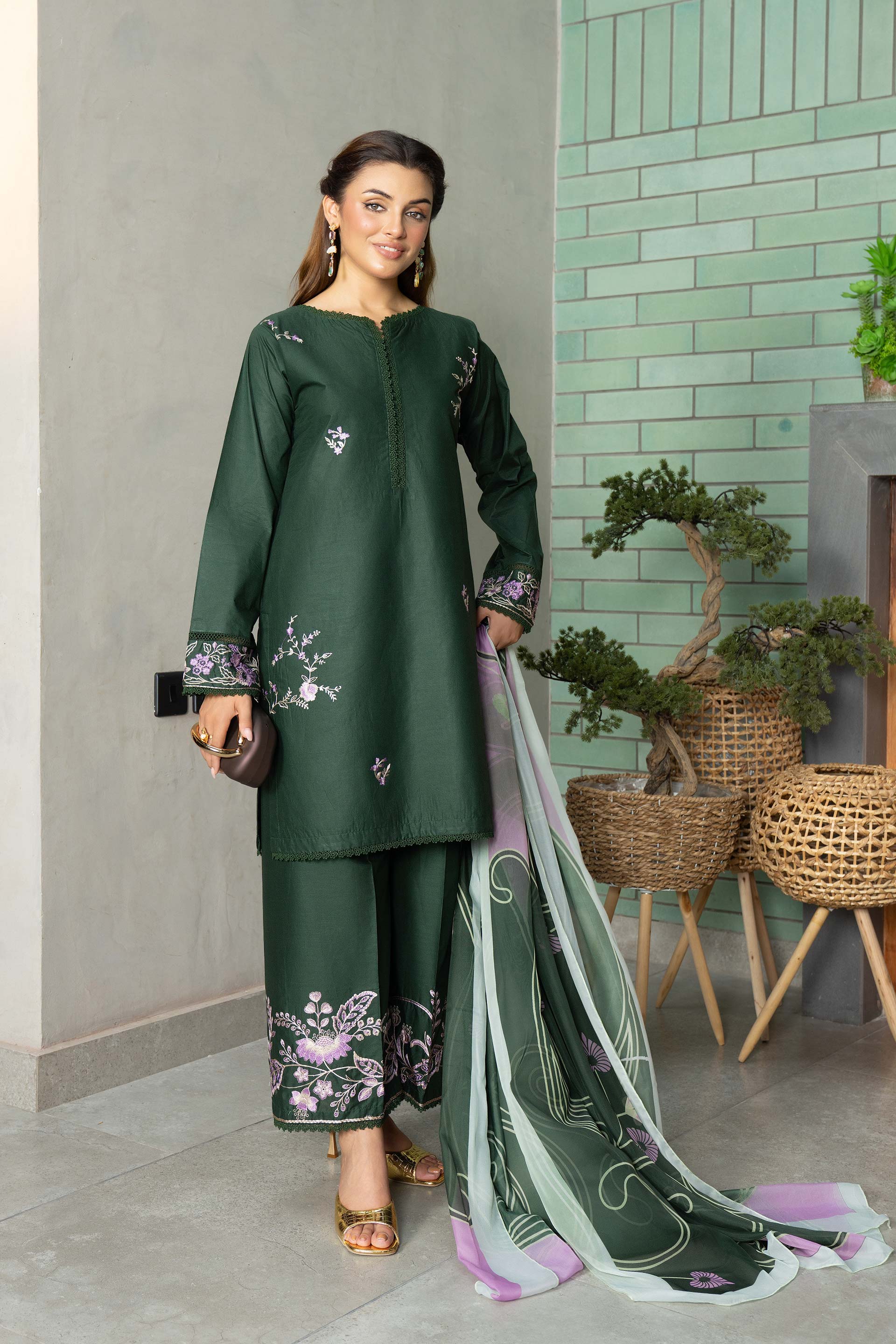 Woman in a green traditional outfit with floral patterns, holding a dupatta, against a light green tiled wall.